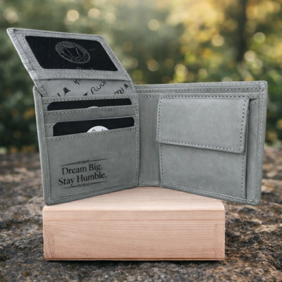 ארנק עור אפור פתוח עם חריטת Dream Big Stay Humble על עור פנימי, מונח על קופסת עץ על רקע טבע מטושטש