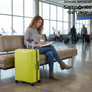 אישה יושבת בשדה תעופה לצד מזוודת סמסונייט (Samsonite) קשיחה בצבע צהוב ליים.